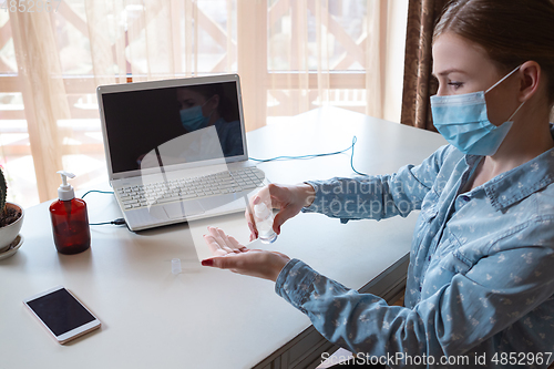 Image of Young woman in face mask disinfecting gadgets surfaces on her workplace