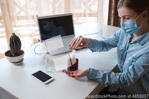 Image of Young woman in face mask disinfecting gadgets surfaces on her workplace