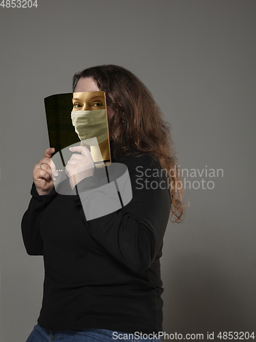 Image of Be safe and read to become someone else - woman covering face with book in face mask while reading on grey background