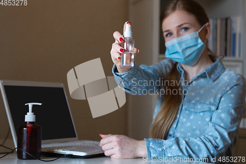 Image of Young woman in face mask disinfecting gadgets surfaces on her workplace