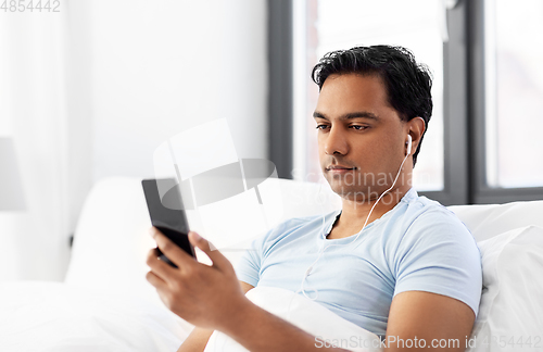 Image of indian man in earphones with phone in bed at home