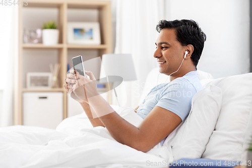 Image of indian man in earphones with phone in bed at home