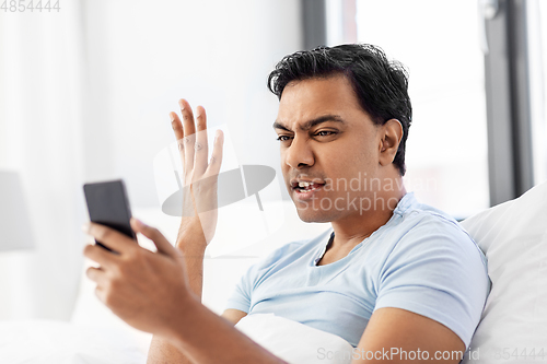 Image of angry indian man with smartphone in bed at home