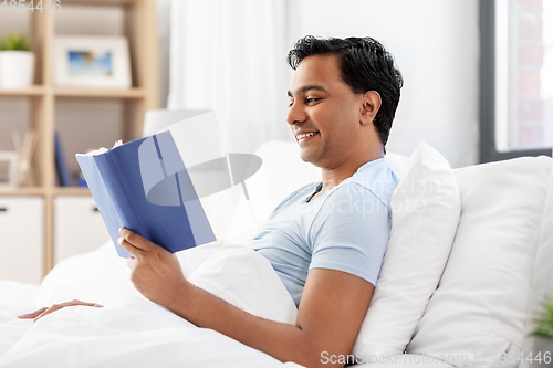 Image of happy indian man reading book in bed at home