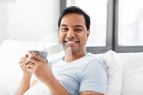 Image of happy indian man drinking coffee in bed at home
