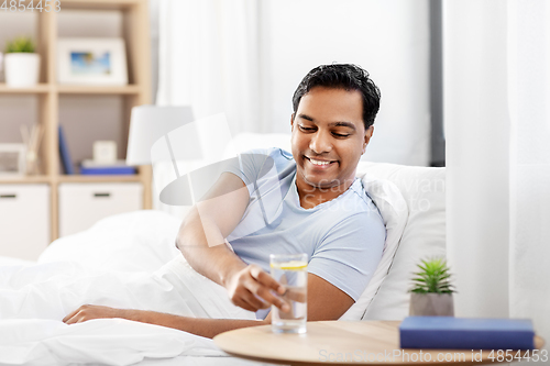 Image of happy indian man drinking water lying in bed