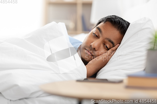 Image of sleepy indian man in bed looking at smartphone