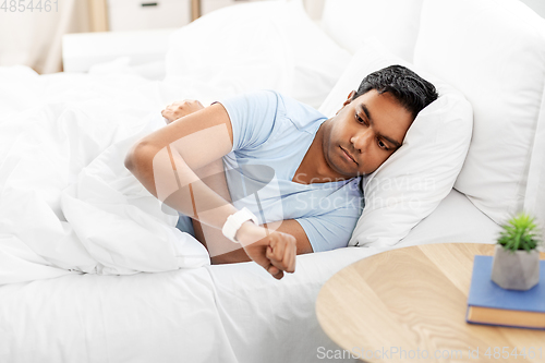 Image of indian man looking at smart watch in bed at home