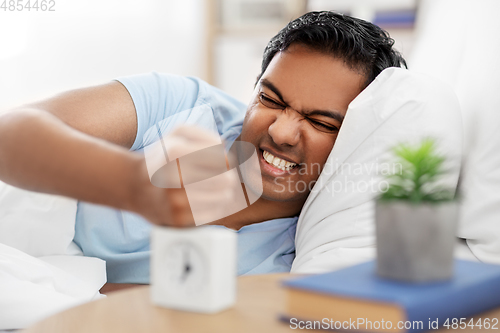 Image of angry indian man with alarm clock lying in bed