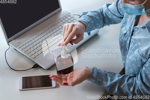 Image of Young woman in face mask disinfecting gadgets surfaces on her workplace