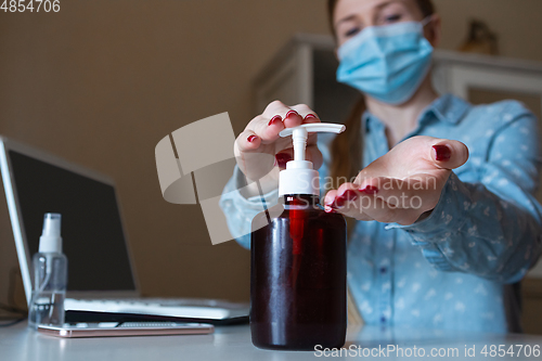 Image of Young woman in face mask disinfecting gadgets surfaces on her workplace