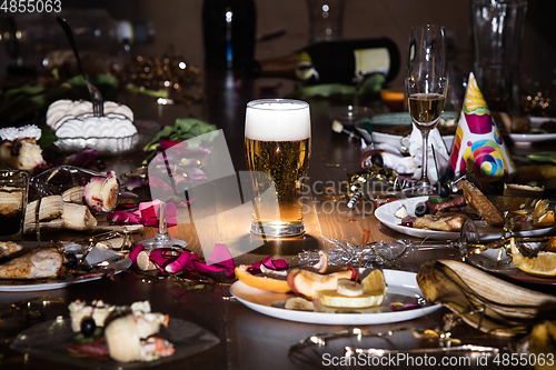 Image of Early morning after the party. Glass of light, cold lager, beer on the table with confetti and serpentine, leftovers, flower petals