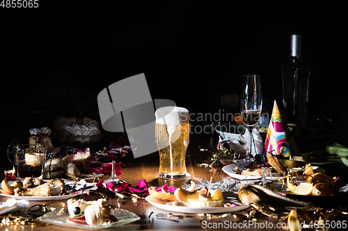 Image of Early morning after the party. Glass of light, cold lager, beer on the table with confetti and serpentine, leftovers, flower petals