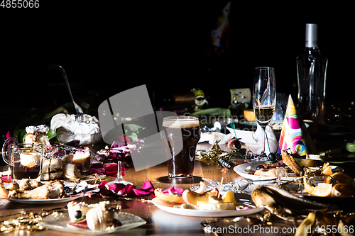 Image of Early morning after the party. Glass of dark, cold stout, beer on the table with confetti and serpentine, leftovers, flower petals