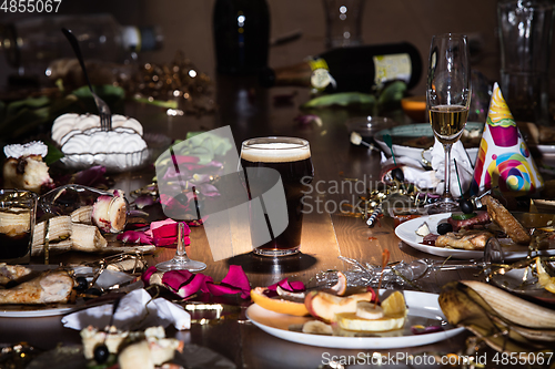 Image of Early morning after the party. Glass of dark, cold stout, beer on the table with confetti and serpentine, leftovers, flower petals