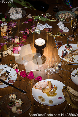 Image of Early morning after the party. Glass of dark, cold stout, beer on the table with confetti and serpentine, leftovers, flower petals