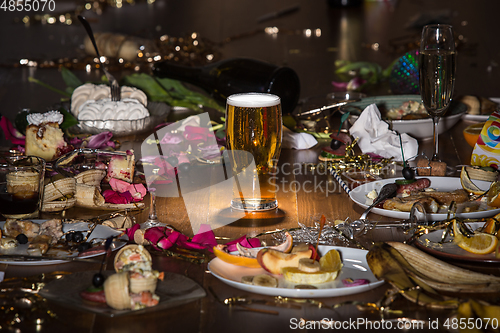 Image of Early morning after the party. Glass of light, cold lager, beer on the table with confetti and serpentine, leftovers, flower petals