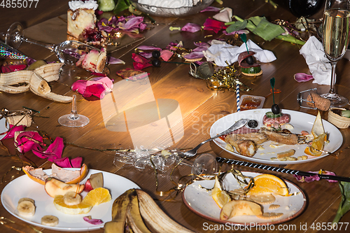 Image of Early morning after the party. Glasses and plates on the table with confetti and serpentine, leftovers, flower petals