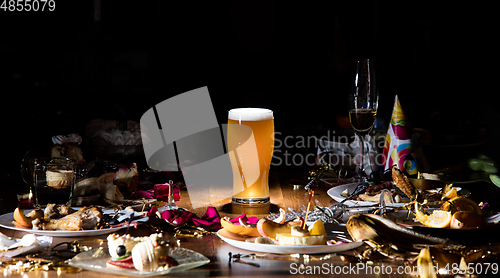 Image of Early morning after the party. Glass of light, cold lager, beer on the table with confetti and serpentine, leftovers, flower petals