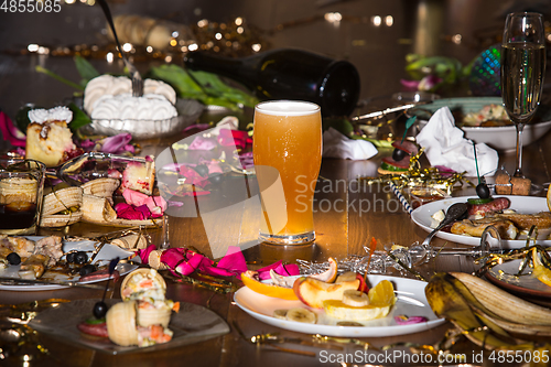 Image of Early morning after the party. Glass of light, cold lager, beer on the table with confetti and serpentine, leftovers, flower petals
