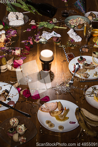 Image of Early morning after the party. Glass of dark, cold stout, beer on the table with confetti and serpentine, leftovers, flower petals