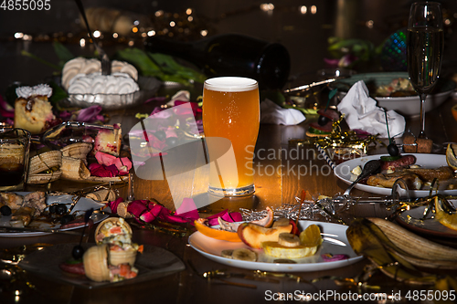 Image of Early morning after the party. Glass of light, cold lager, beer on the table with confetti and serpentine, leftovers, flower petals