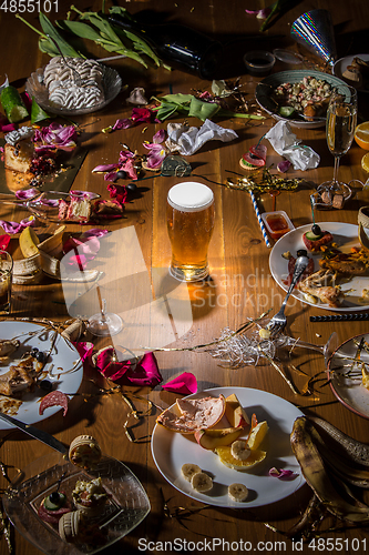 Image of Early morning after the party. Glass of light, cold lager, beer on the table with confetti and serpentine, leftovers, flower petals