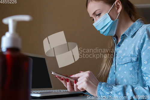 Image of Young woman in face mask disinfecting gadgets surfaces on her workplace