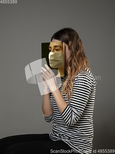 Image of Be safe and read to become someone else - woman covering face with book in face mask while reading on grey background