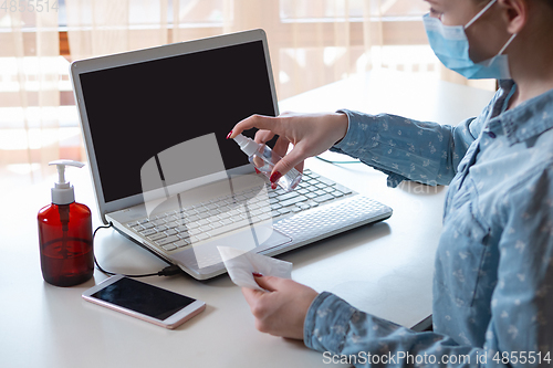 Image of Young woman in face mask disinfecting gadgets surfaces on her workplace