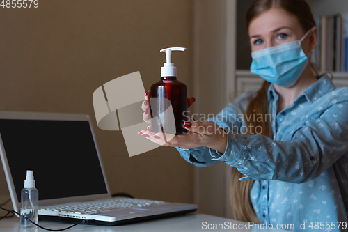 Image of Young woman in face mask disinfecting gadgets surfaces on her workplace