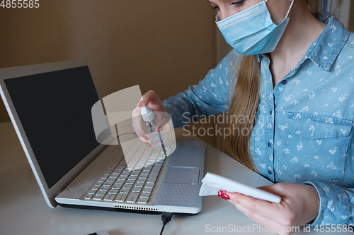 Image of Young woman in face mask disinfecting gadgets surfaces on her workplace