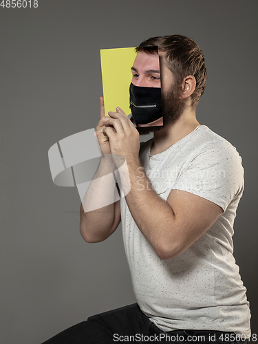 Image of Be safe and read to become someone else - man covering face with book in face mask while reading on grey background