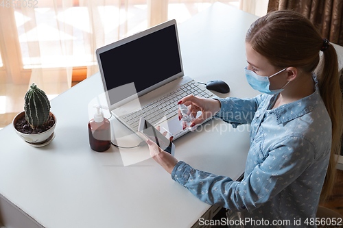 Image of Young woman in face mask disinfecting gadgets surfaces on her workplace