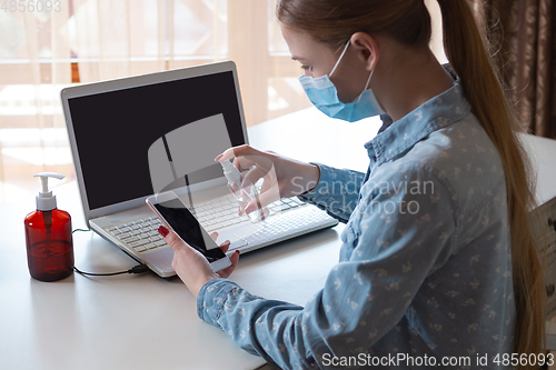 Image of Young woman in face mask disinfecting gadgets surfaces on her workplace