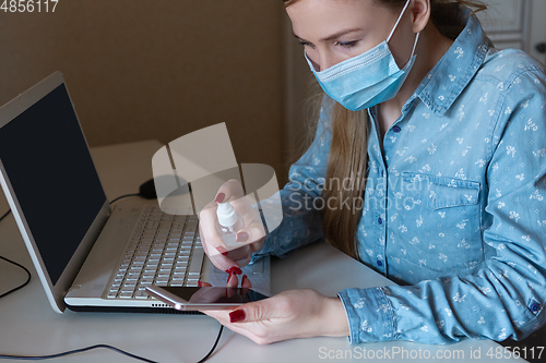 Image of Young woman in face mask disinfecting gadgets surfaces on her workplace