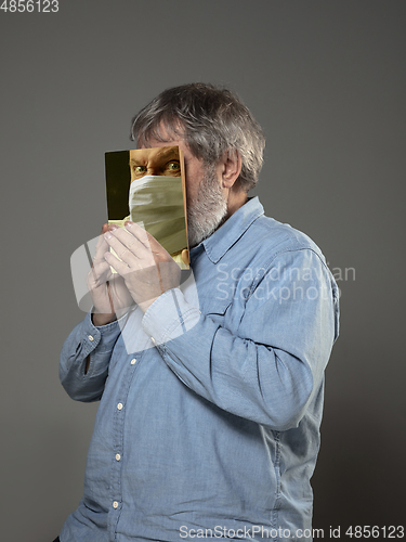 Image of Be safe and read to become someone else - man covering face with book in face mask while reading on grey background