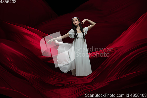 Image of Young and graceful ballet dancer on billowing red cloth background in classic action. Art, motion, action, flexibility, inspiration concept.