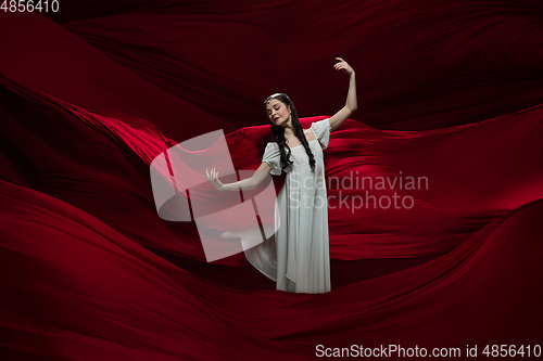 Image of Young and graceful ballet dancer on billowing red cloth background in classic action. Art, motion, action, flexibility, inspiration concept.