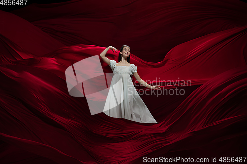 Image of Young and graceful ballet dancer on billowing red cloth background in classic action. Art, motion, action, flexibility, inspiration concept.