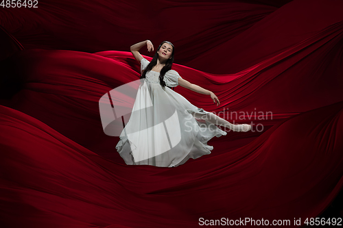 Image of Young and graceful ballet dancer on billowing red cloth background in classic action. Art, motion, action, flexibility, inspiration concept.