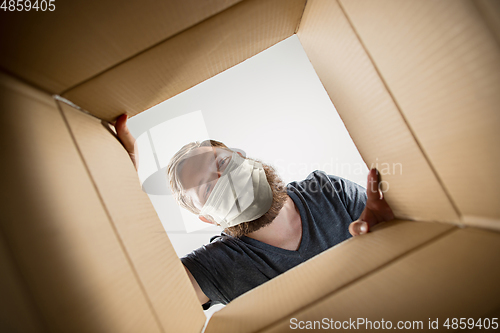 Image of Young man opening the huge postal package wearing protective face mask, contactless delivery