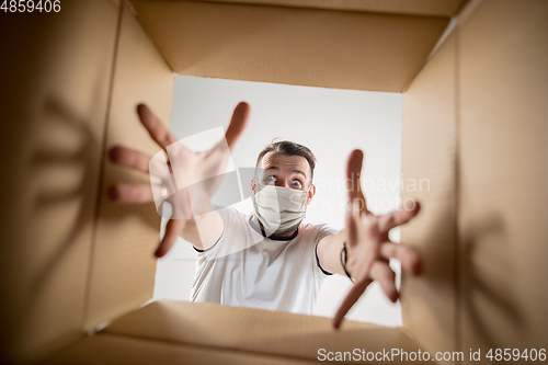 Image of Young man opening the huge postal package wearing protective face mask, contactless delivery