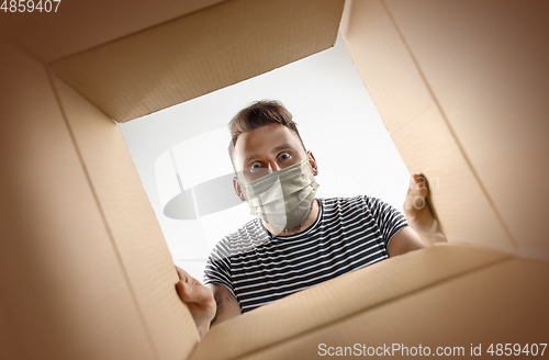 Image of Young man opening the huge postal package wearing protective face mask, contactless delivery