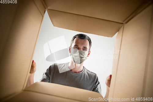 Image of Young man opening the huge postal package wearing protective face mask, contactless delivery