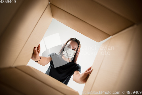 Image of Young woman opening the huge postal package wearing protective face mask, contactless delivery