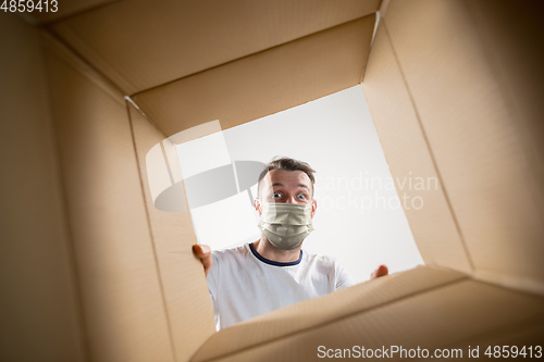 Image of Young man opening the huge postal package wearing protective face mask, contactless delivery