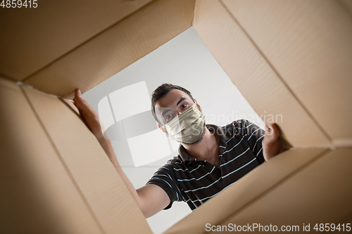 Image of Young man opening the huge postal package wearing protective face mask, contactless delivery