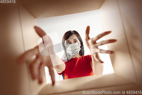 Image of Young woman opening the huge postal package wearing protective face mask, contactless delivery