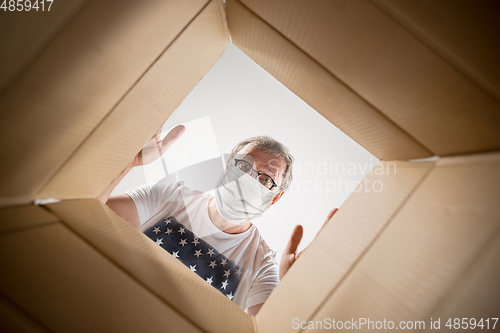 Image of Senior man opening the huge postal package wearing protective face mask, contactless delivery